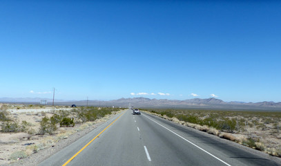 Naklejka premium Autobahn in der Mojave-Wüste/Lange, gerade Autobahn durch die Mojave-Wüste in Kalifornien, zweispurige Fahrbahn mit Markierung und Standstreifen, bergige Landschaft und blauer Himmel