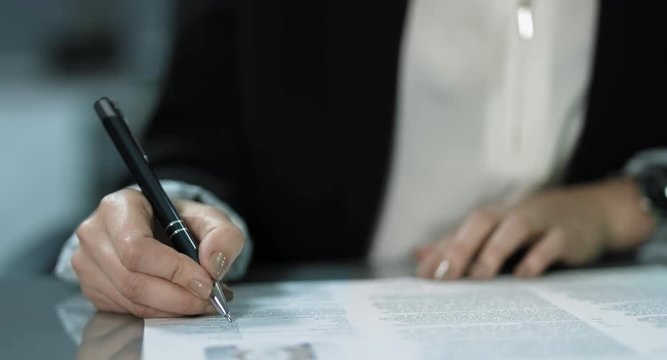 4K: Ein junge Geschaeftsfrau unterzeichnet einen Vertrag an ihrem Schreibtisch. Die Frau tr&auml;gt eine wei&szlig;e Bluse mit einer schwarzen Jacke. Ihre N&auml;gel sind dezent lackiert.