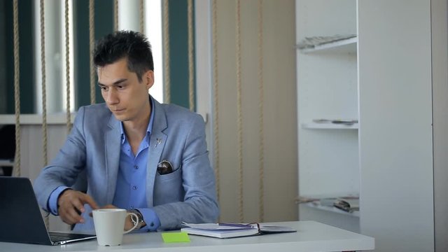 Male is taking coffee break sitting at workplace continuing working.