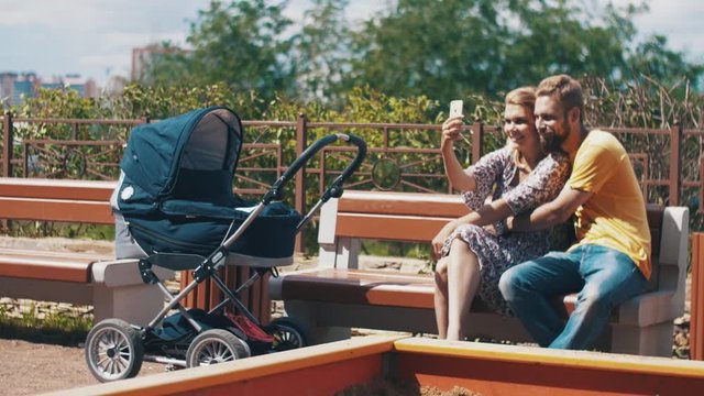 Young parents with baby carriage on bench. Take selfie on smartphone. Family. Playground. Summer sunny day