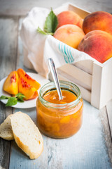 A jar with homemade peach jam in front of a white wood crate full of ripe peaches over a light blue old wood background.