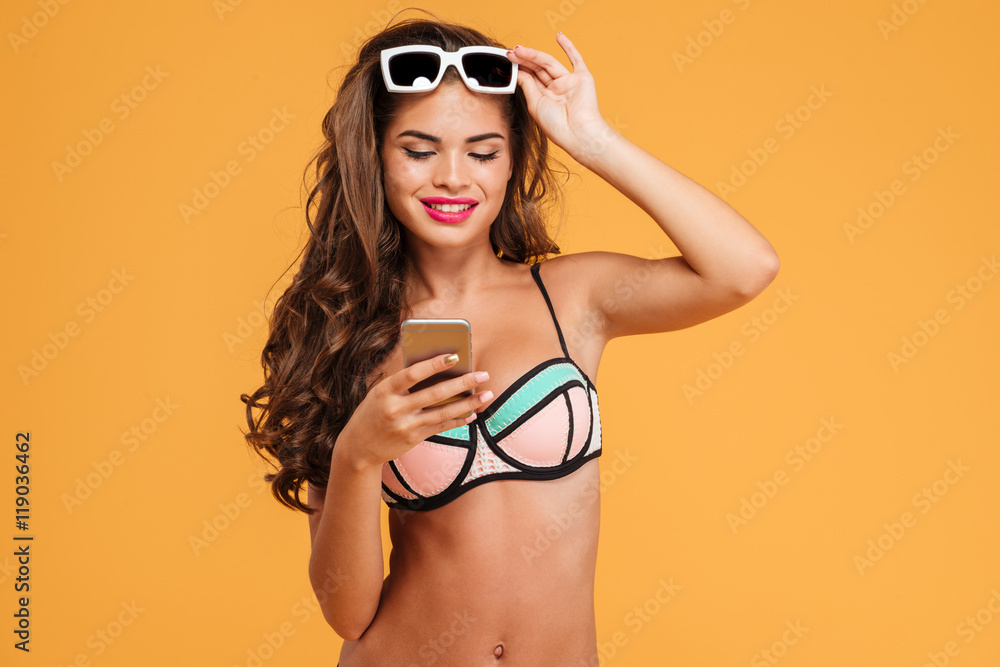 Wall mural Young sexy girl in bikini using smartphone and holding suglasses