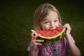 Cute little blond girl eating watermelon on the grass in summertime