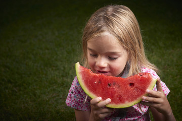 Cute little blond girl eating watermelon on the grass in summertime