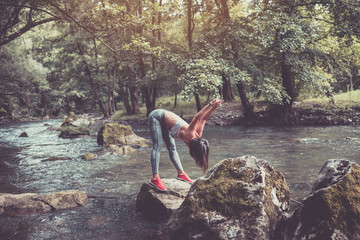 Attractive young woman doing stretching exercises in beautiful untouched nature environment. She standing on the huge rock in the middle of cold mountain river.