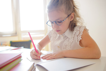 Cute little schoolgirl in glasses something diligently writes in a notebook.