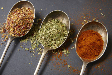 Three antique silver spoons with assorted cooking seasonings arranged on a dark slate background