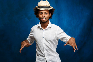 Young handsome african man in hat dancing over blue background.