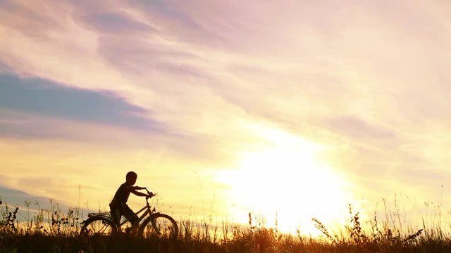 9 yeard old boy riding bike then celebrating his success with funy dance. Silhouette of little jumping happy kid with hands up at sunset sky background. Real time bideo footage.