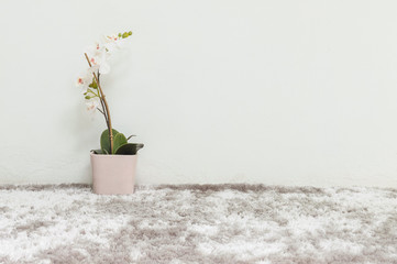 Closeup artificial plant with white orchid flower in pink pot on blurred gray carpet and white cement wall textured background under window light