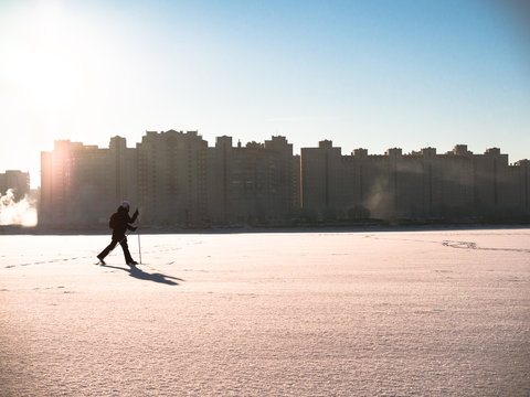 Toned image of an adult woman and skiing with a backpack walking on snow against the backdrop of urban homes and clear sky with sun