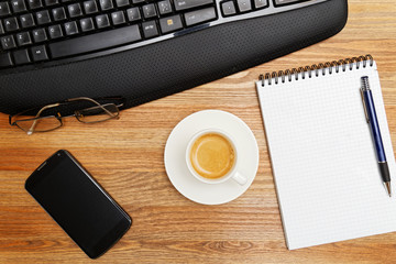 Wooden desktop with a cup of coffee in the center. Near blank notebook, pen, smartphone, glasses and keyboard. Top view. Copyspace.