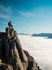 Gordijnen Adult woman with a backpack and Alpenstocks stands on the edge of a cliff and looking at the sunrise against the blue sky and thick clouds floating down © atomfotolia