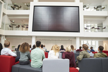 Students watching big screen in university atrium, back view