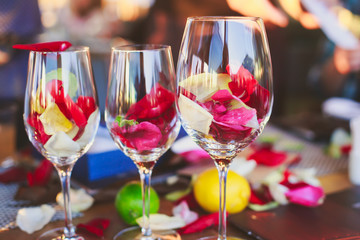 Beautiful glass with rose leafs, rose-petal, rose petals on a romantic date with wine, candles in candlelight and with celebrating couple on the background, interior for a romantic date diner 