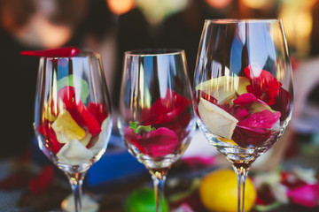 Beautiful glass with rose leafs, rose-petal, rose petals on a romantic date with wine, candles in candlelight and with celebrating couple on the background, interior for a romantic date diner 