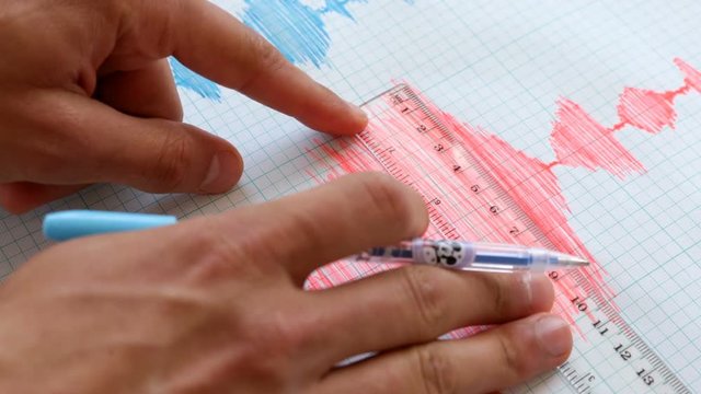 Seismological device for measuring earthquakes. Seismological activity lines on the sheet of measuring paper. Earthquake wave on graph paper. Human hand writing down with a blue pen.