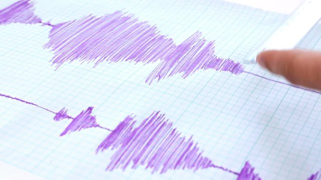 Seismological device for measuring earthquakes. Seismological activity lines on the sheet of measuring paper. Earthquake wave on graph paper. Human hand writing down with a blue pen.