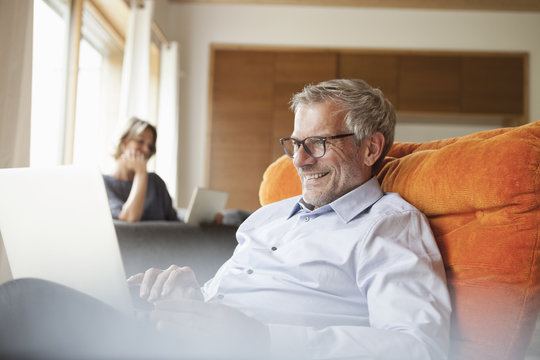 Smiling man using laptop in armchair with wife in background