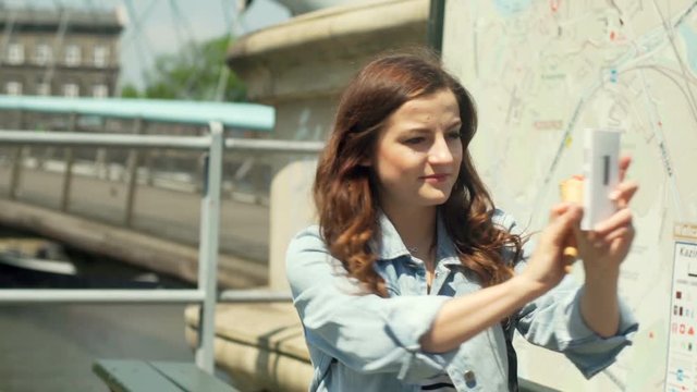 Pretty girl standing with ice cream next to the map and doing selfies on smartphone
