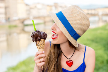 Young female tourist in blue dress holding chocolate ice cream in waffle cone sitting near the river in Florence city. Eating traditional italian ice cream gelato.