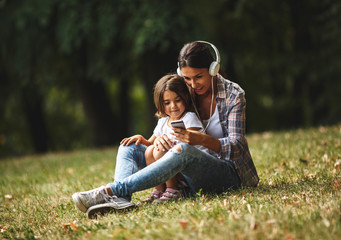 Mother and daughter sitting on grass and listening to music on smart phone.