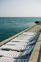 Fototapeta premium View of line of white deckchairs on cement coast against the sea.copy space
