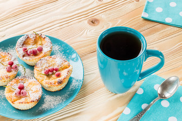 cheesecakes decorated frozen red currant berries and powdered sugar on blue plate, black tea in blue cup, blue napkin at white polka dots, old silver spoon  on wooden table