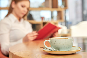 Fototapeta premium Break from work. Selective focus on a cup on the table business woman reading a book on the background