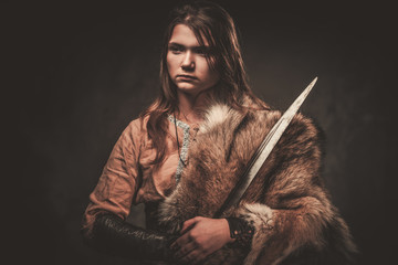 Serious viking woman with sword in a traditional warrior clothes, posing on a dark background.