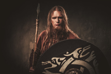 Serious viking with a spear in a traditional warrior clothes, posing on a dark background.