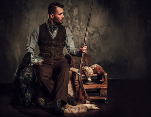 Handsome hunter with a english setter and shotgun in a traditional shooting clothing, sitting on a...