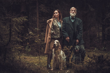 Two hunters with dogs and shotguns in a traditional shooting clothing, posing on a dark forest...