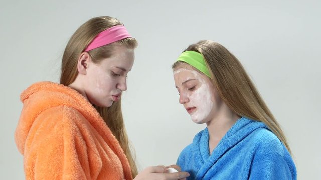 Teenage girls in bathrobes with applied homemade facial cream masks girl using cotton pad wiping off mask on the face of her girlfriend