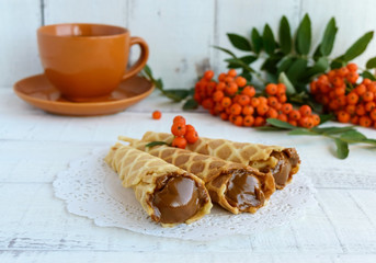 Fototapeta na wymiar Homemade honey wafers rolled into a cone, filled with caramel (condensed milk), a cup of tea, rowan berries on a white background. Close up