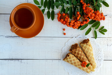 Homemade honey wafers rolled into a cone, filled with caramel (condensed milk), a cup of tea, rowan berries on a white background. top view