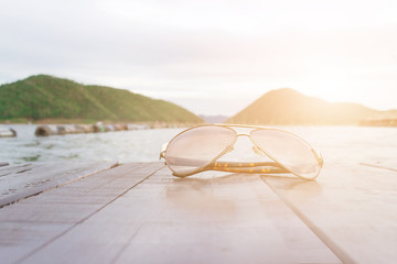 Fototapeta na wymiar Sunglasses Placed on a wooden floor and Mountain river sky sun