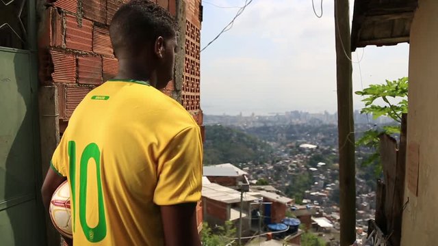 A kid in a soccer jersey stands with a ball under his arm while looking out at the city of Rio de Janeiro