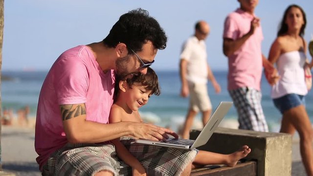 A father and son sit by the beach and look at a laptop