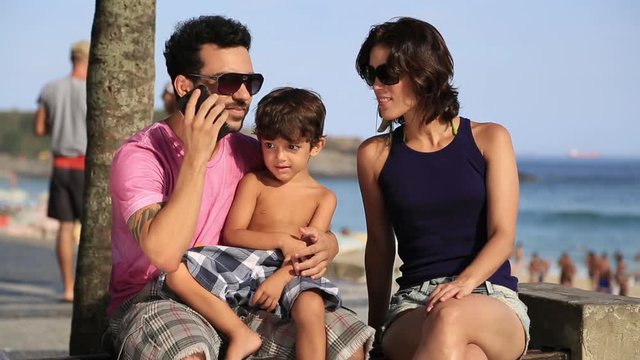 A young family sits near the beach while the father talks on his smart phone