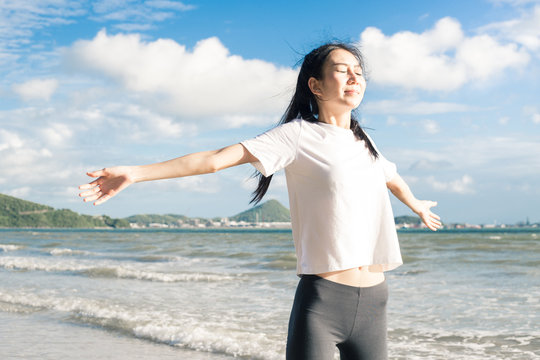 Happy Relaxed Asian Woman Breathing Deep Fresh Air And Raising Arms On The Beach With Blue Sky Background