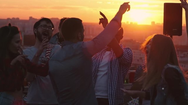 Group of happy young multi-ethnic friends dancing energetically and raising their arms up at sunset rooftop party