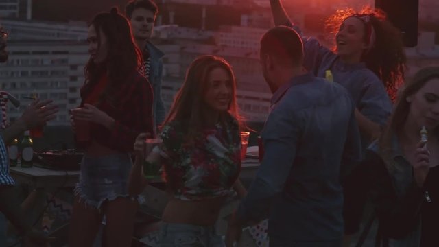 Group of young multi-ethnic people enjoying spending time at rooftop sunset party, dancing to the music played by dj, drinking cocktails and smoking hookah, beautiful couple kissing