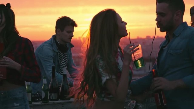 Medium shot of beautiful young couple dancing at sunset rooftop party to the music played by dj, drinking cocktails and beer surrounded by friends