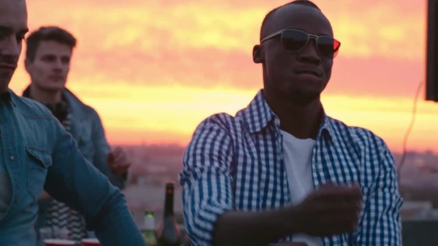 Medium shot of African American young man in sunglasses dancing at sunset rooftop party to the music played by dj