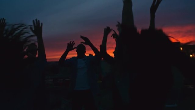 Silhouettes of group of young people dancing and raising their arms up in the air against beautiful sunset sky at rooftop party 