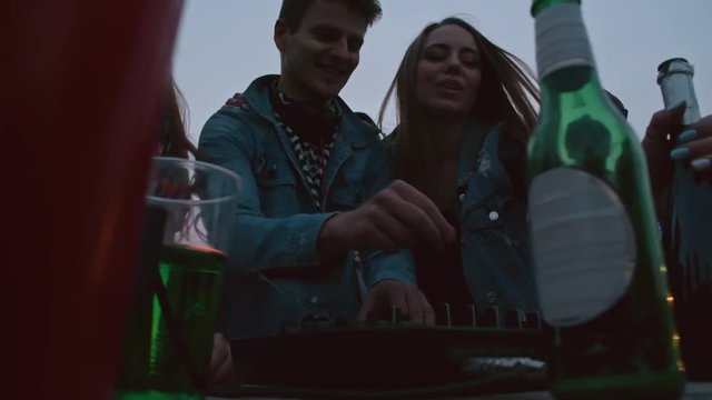 Young dj playing music on rooftop surrounded by group of beautiful multi-ethnic young people at sunset, beer bottles and cocktails near turntable