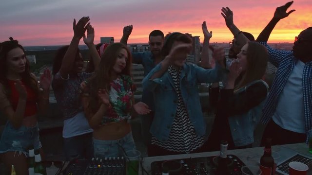 Young dj playing music on rooftop surrounded by group of multi-ethnic young people dancing and clapping hands at sunset, beer bottles and cocktails near turntable