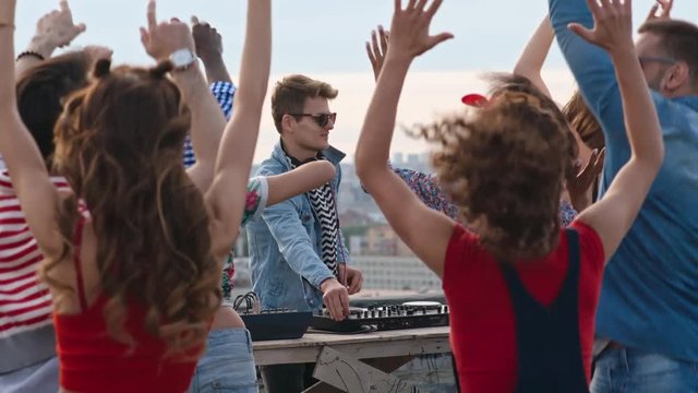 Dj mixing music at rooftop day party surrounded by crowd of multi-ethnic young clubbers dancing, jumping and waving their arms in the air 