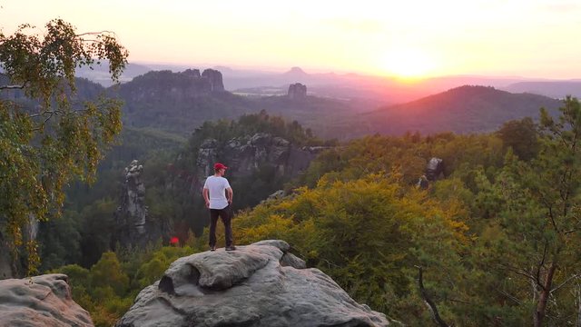 Tourist in grey shirt and trekking trousers photograph. Man takes photos or record video with smart phone on peak of rock empire park. Summer sunset above beautiful valley of Saxony Switzerland park. 